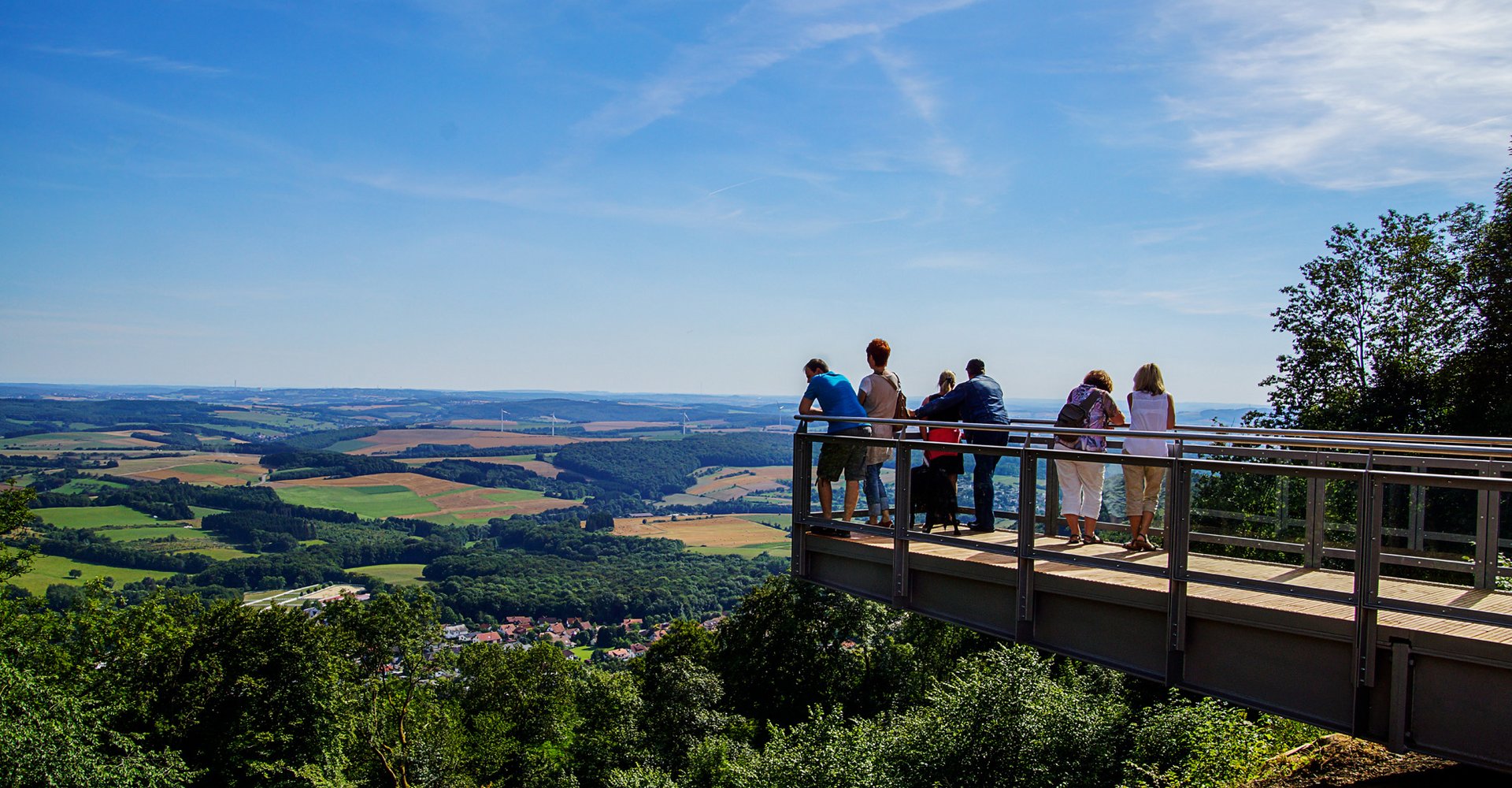 Personen stehen auf dem Aussichtssteg auf dem Schaumberg und genießen den Blick auf das Sankt Wendeler Land.
