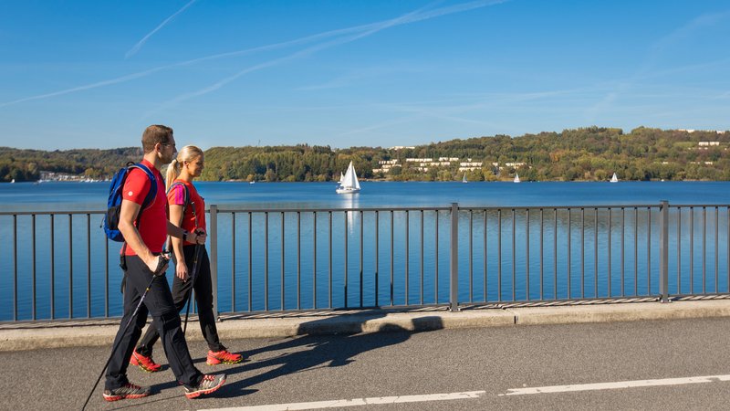 Eine Frau und ein Mann wandern, bepackt mit einem Rucksack und mit Wanderstöcken in der Hand, über den Staudamm des Bostalsees: Im Hintergrund sind Segelboote auf dem Wasser zu sehen.