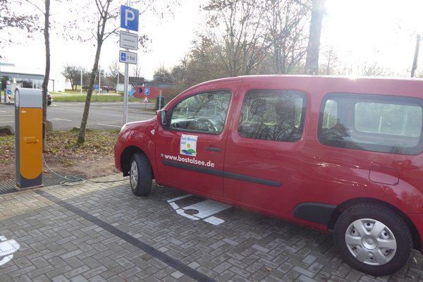 Ein rotes Auto mit der Aufschrift www.bostalse.de steht auf einem Parkplatz an einer Ladesäule für Elektroautos und lädt über einen Schlauch zur Ladesäule seinen Akku auf.