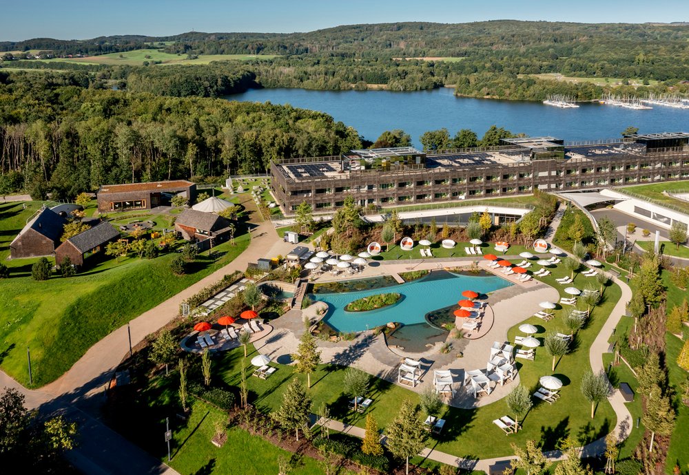 Seezeit entdecken In einer Luftaufnahme des Wellnesshotels Seezeitlodge ist das Gebäude des Hotels, das keltische Saunadrof sowie der Außenschimmbereich mit Sonnenliegen und Bar zu sehen. Die Umgebung ist in eine grüne Landschaft eingebettet.