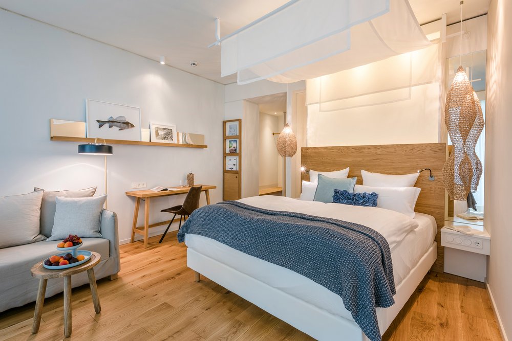Seezeit Träumereien Es ist ein helles Hotelzimmer des Wellnesshotels Seezeitlodge mit warmen Blautönen, ausgestattet mit einem Bett, Sofa, Coutisch und Regal und Nachttischen zu sehen.