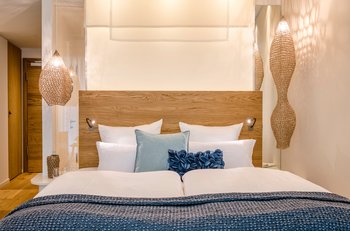 Ein gemütlich beleuchtetes Zimmer mit einem großen Doppelbett mit einer blauen Tagesdecke und vielen Kissen darauf.