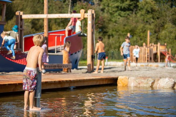 Wasserspielplatz Ein Junge steht im Wasser auf dem Wasserspielplatz und spritzt Wasser aus einer Standpumpe. Dahinter spielen Kinder auf einem blau-roten Schiff oder im Sand.