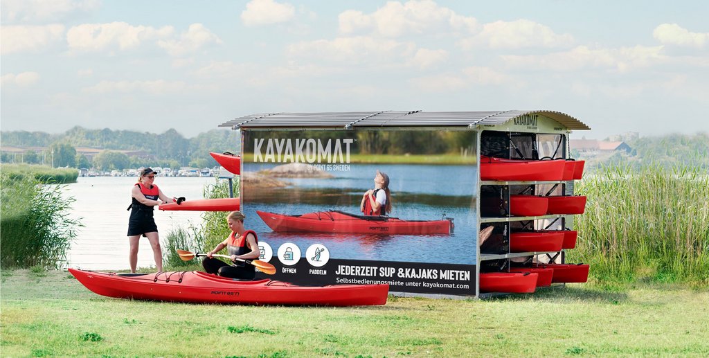 Foto: Malte Danielsson AB Eine Kajak Verleihstation an einem See. In einem überdachten Gehäuse sind mehrere Kajaks sowie SUP-Boards gelagert. Eine Frau ist vor einem auf dem Boden liegenden Kajak in der Hocke und baut ein Paddel zusammen und eine andere Frau zieht ein Kajak aus der Verleihstation.