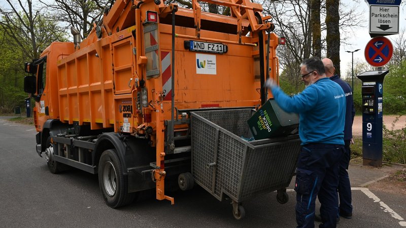 Zwei Männer stehen an einem orangenen Müllauto und leeren einen Mülleimer in den Fangkorb, der hinten am Fahrzeug hängt.