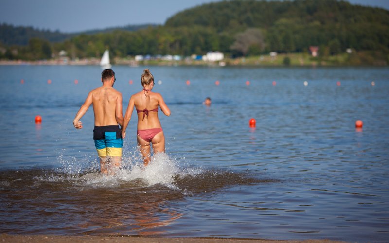 Ein junges Paar ist von hinten zu sehen, während es Hand in Hand in den Badesee Bostalsee läuft.