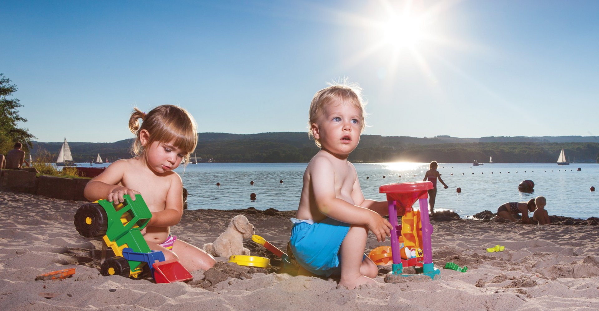 2 Kleinkinder, ein Junge und ein Mädchen, spielen im Strandbad mit ihrem bunten Spielzeug im Sand