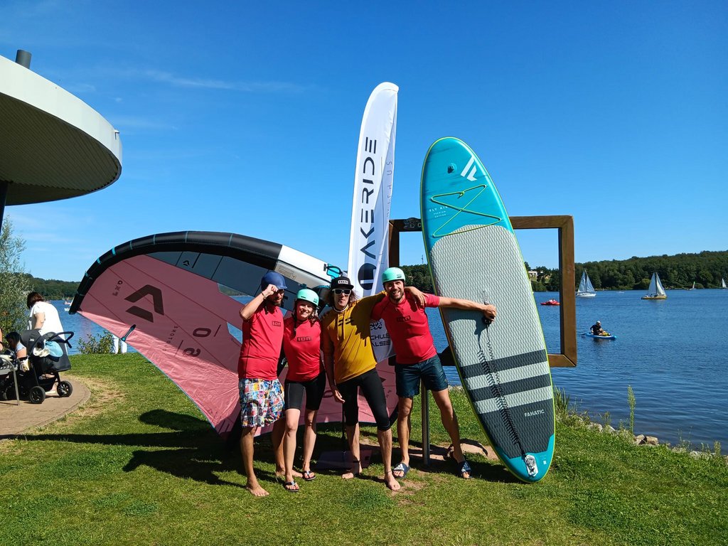 Photo: Lakeride 4 Personen stehen am Ufer des Bostalsees und haben ein Wingfoil, ein SUP und eine Beachflag mit dabei.