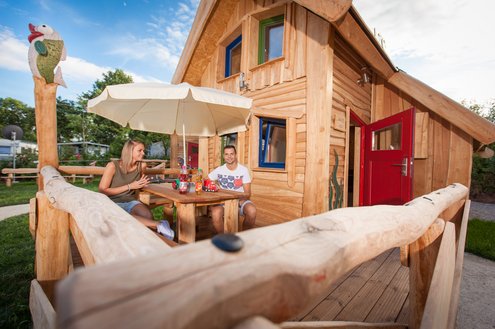 Im Fokus des Fotos befindet sich ein Märchenhaus. Es ist ein kleines Holzhaus, mit einer Terrasse und Spielgerät. Auf der Terrasse sitzt ein junges Elternpaar bei einem Kaffee.