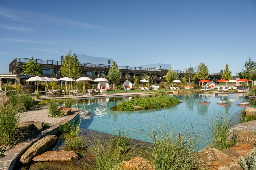 Seezeit zu Zweit Es ist die Außenanlage des Wellnesshotels Seezeitlodge zu sehen. In der Umgebung sieht man Sonnenliegen, eine Bar und einen Schwimmteich.