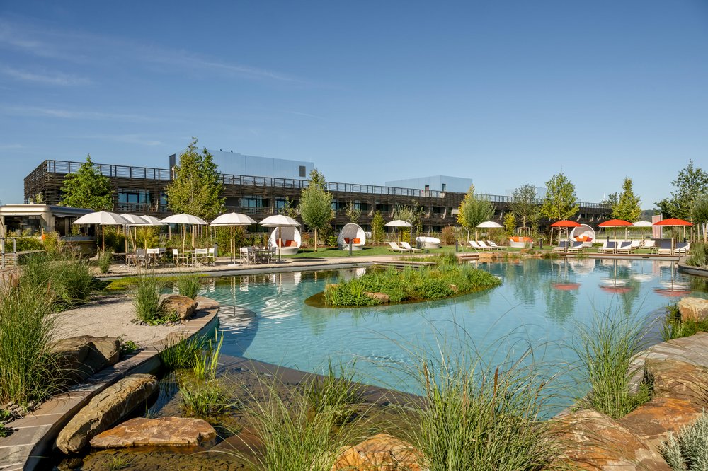 Es ist die Außenanlage des Wellnesshotels Seezeitlodge zu sehen. In der Umgebung sieht man Sonnenliegen, eine Bar und einen Schwimmteich. 