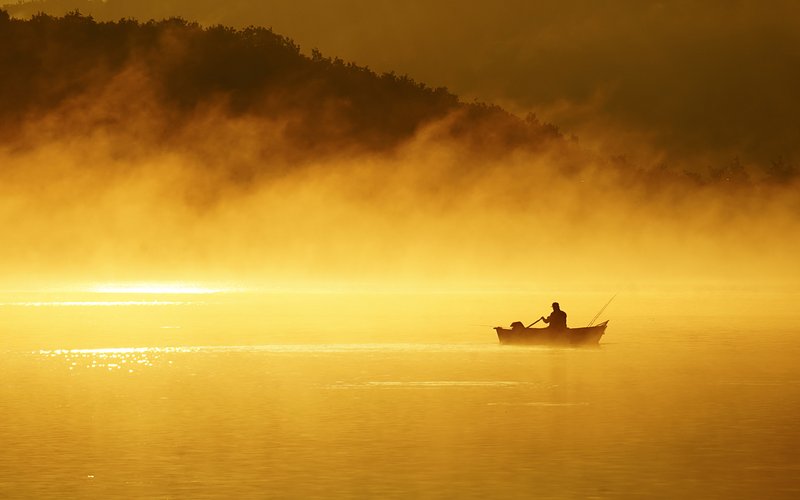 Bei Sonnenaufgang ist ein Angler in seinem Ruderboot mitten auf einem See zu sehen. Der See liegt in goldenem Licht.