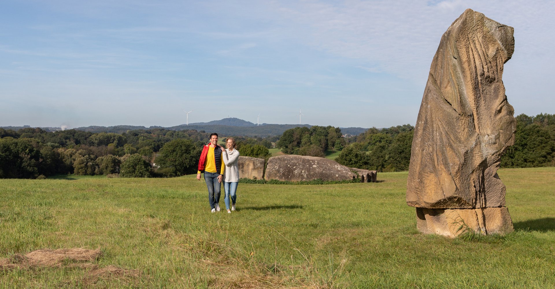 Ein Mann und eine Frau laufen über eine Wiese. Hinter und neben ihnen befinden sich große Steinskulpturen.