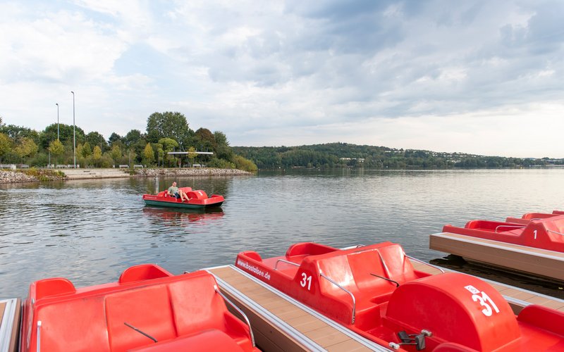 Am Steg des Bootsverleih Bosen weilen in Reih und Glied rotfarbene Tretboote, während in der Ferne ein junger Mann mit einem roten Tretboot auf dem Wasser unterwegs ist