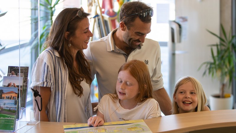 Eine Familie mit zwei Mädchen steht an der Theke der Rezeption des Campinglatzes. Das größere Mädchen zeigt auf etwas auf dem Lageplan des Campingplatzes.