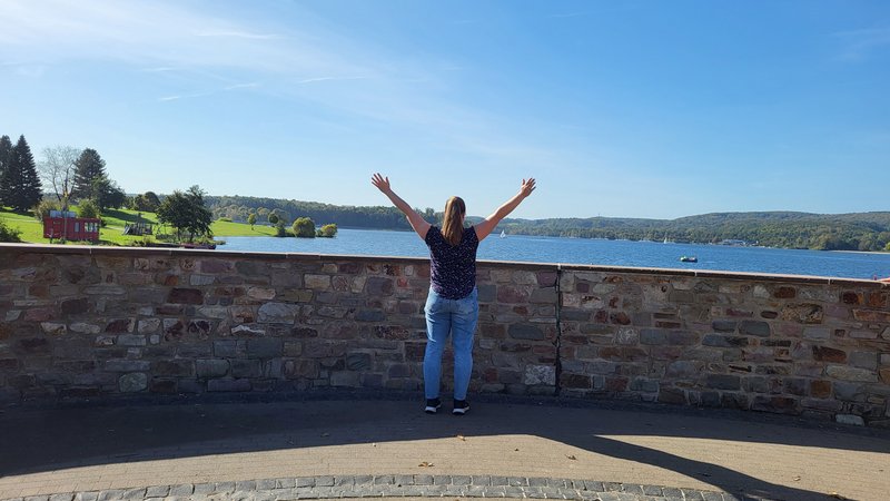 Eine junge Frau, mit dem Blick abgewandt, steht an der Seekanzel des Bostalsees und schaut aufs Wasser. Dabei hat streckt sie die Arme in die Luft