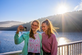 Zusammen stark Zwei junge Frauen stehen auf dem Staudamm der Talsperre Nonnweiler. Im Hintergrund ist die Natur und das Wasser zu sehen. Sie selbst stehen im Vordergrund, eine hält ein Smartphone, mit dem sie ein Selfiebild machen.