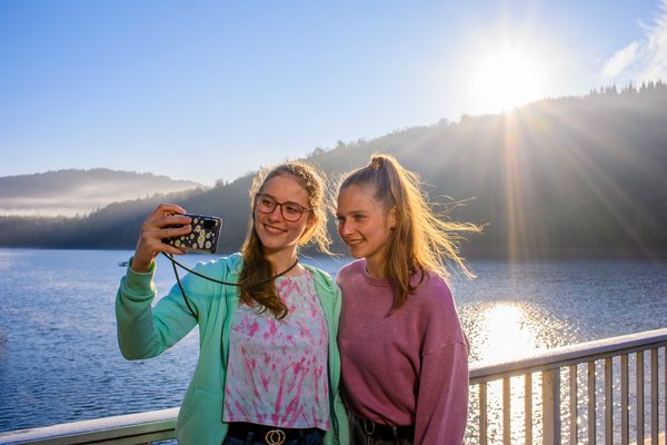 Zusammen stark Zwei junge Frauen stehen auf dem Staudamm der Talsperre Nonnweiler. Im Hintergrund ist die Natur und das Wasser zu sehen. Sie selbst stehen im Vordergrund, eine hält ein Smartphone, mit dem sie ein Selfiebild machen.