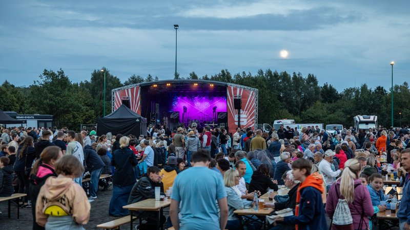 Auf dem Seefest ist viel los. Auf der Festwiese ist eine Bühne aufgebaut, auf der eine Band auftritt. Auf dem Platz selbst sind Bierzeltgarnituren aufgestellt, an denen Menschen sitzen, der Musik zuhören, essen und trinken.