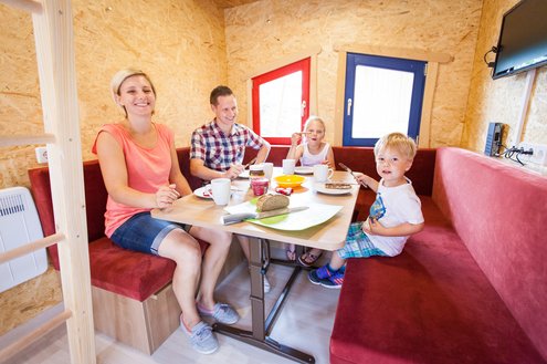 In einem aus Holz gebauten Märchenhäsu sitzt eine Familie mit zwei kleinen Kindern in der Sitzecke und genießt ihr Frückstück.