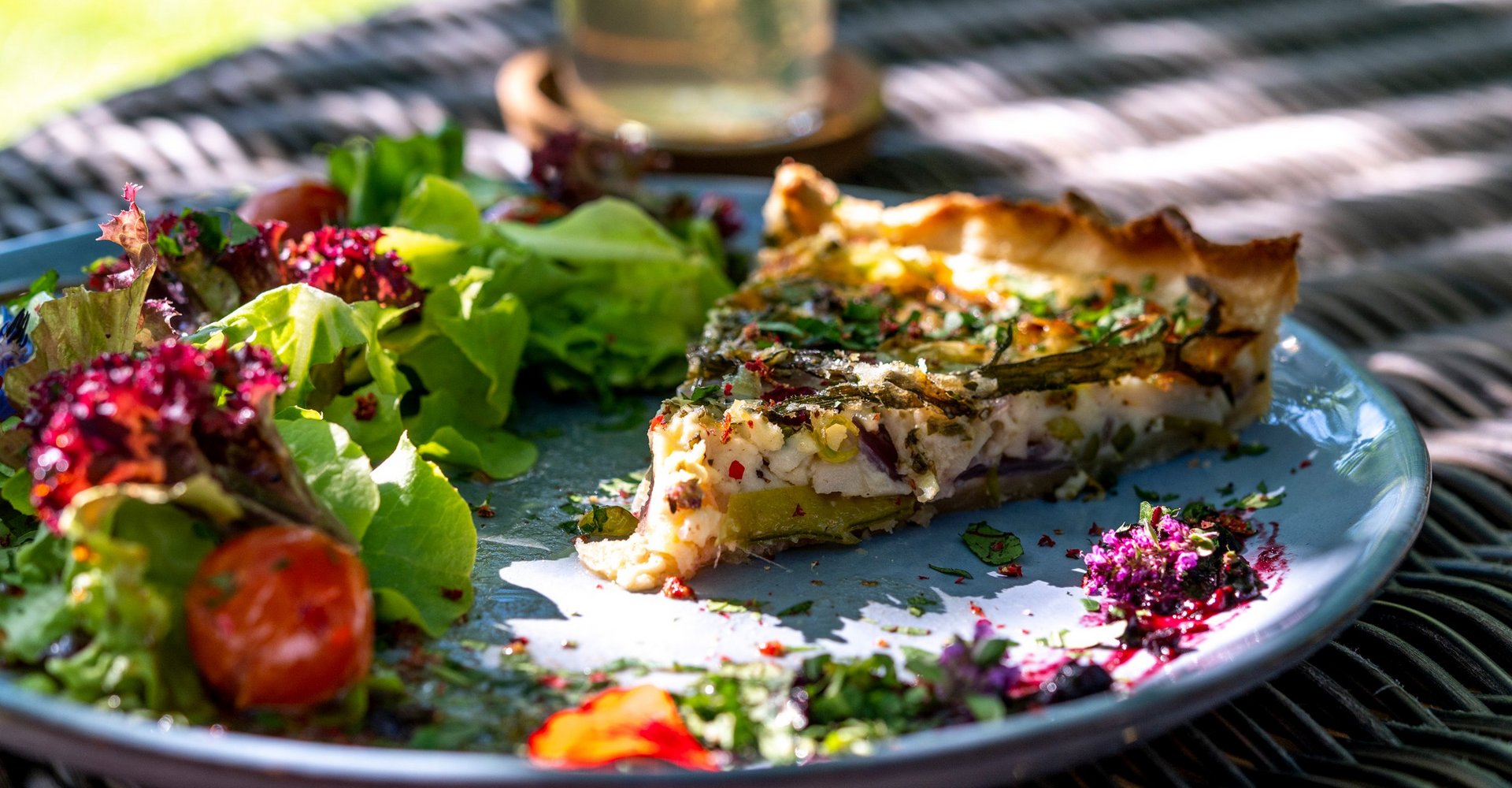 Auf einem Teller mit Salatgarnitur liegt ein Stück Quiche. Der Teller steht im Garten auf einem mit einer Tischdecke gedeckten Tisch.