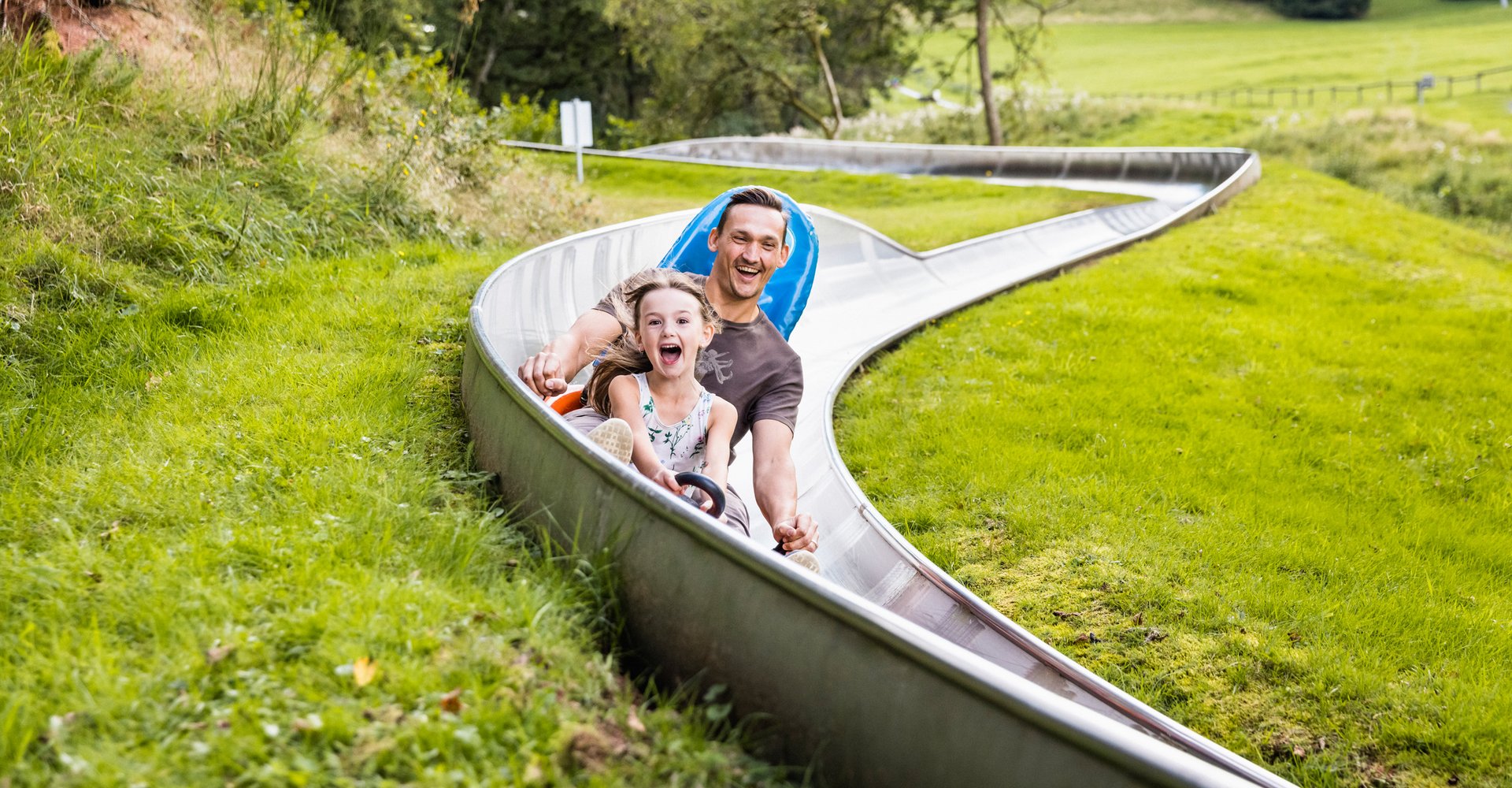 Ein Vater fährt mit seiner Tochter in einem Bob die Sommerrodelbahn hinunter.
