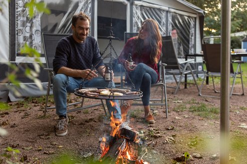 Vor einem Mietwohnwagen des Campingplatzes Bostalsee hat es sich ein Pärchen mit einem Getränk am Grillfeuer, über dem ein Schwenker hängt, gemütlich gemacht.