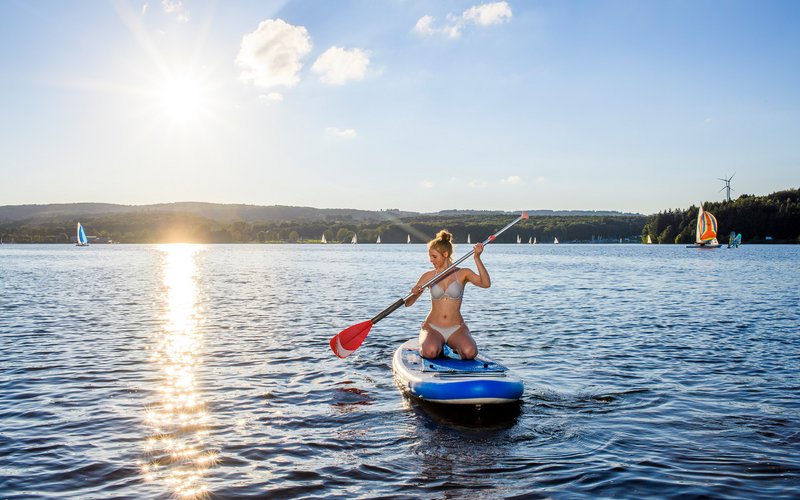 Eine junge Frau kniet Richtung Kamera auf einem Stand Up-paddling Board, das auf dem Bostalsee schwimmt. In ihren Händen hat sie ein zu ihrer rechten ausgerichtetes Paddel, mit dem sie sich vorwärts bewegen möchte 