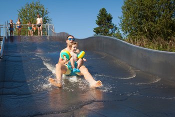 Ein junger Mann rutscht mit Kleinkind in Schwimmflügeln eine wellenförmige Wasserrtusche herunter. Oben, am Rand der Rutsche warten weitere Personen, um ebenfalls herunterrutschen zu können.