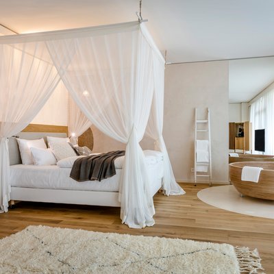 Ein Zimmer mit einem großen Himmelbett mit weißen Vorhängen sowie einem weißen Teppich und einer freistehenden Holzwanne.