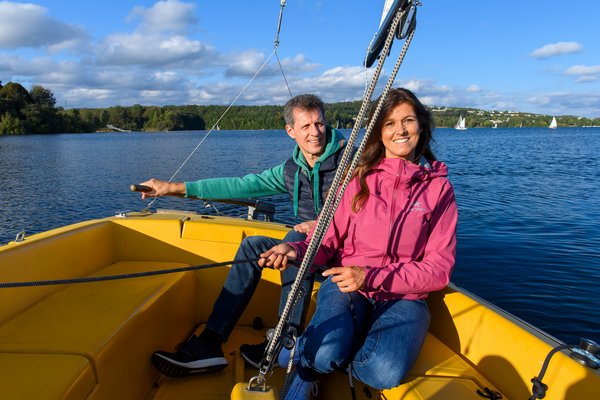 Segeln auf dem Bostalsee Ein Paar mittleren Alters sitzt am Heck eines gelben Segelboots. Die Dame hält die Seile der Segel, während der Herr das Segelboot am Heckruder steuert. Im Hinergrund sind der Bostalsee mit weiteren Segelbooten sowie das Seeufer zu sehen.